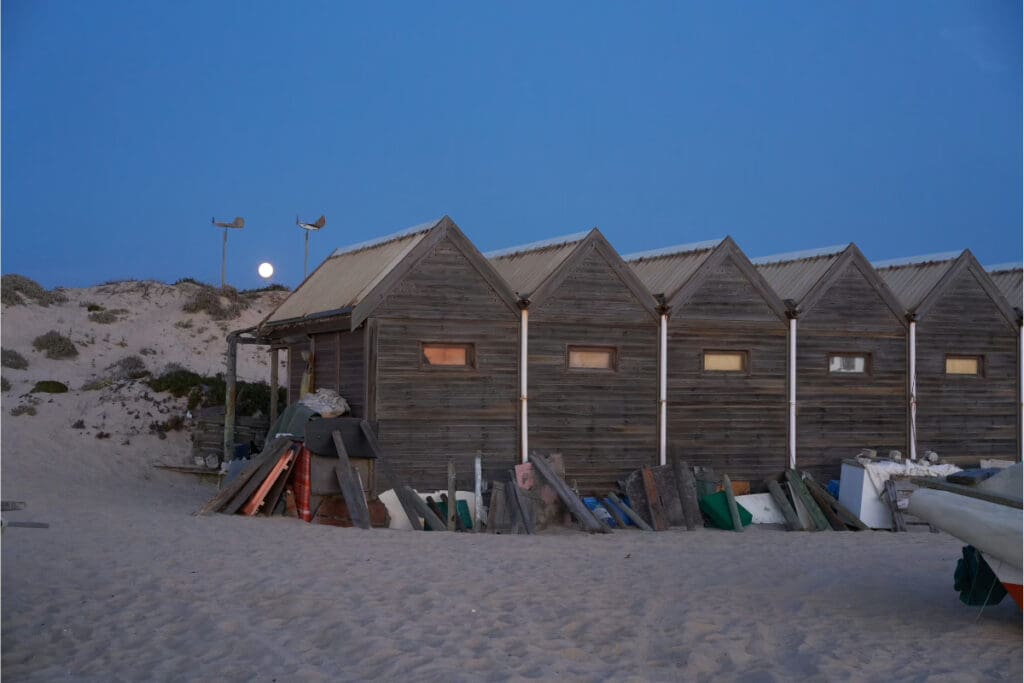 Hidden Places Around the World,Row of wooden beach huts with a full moon in the background at dusk.