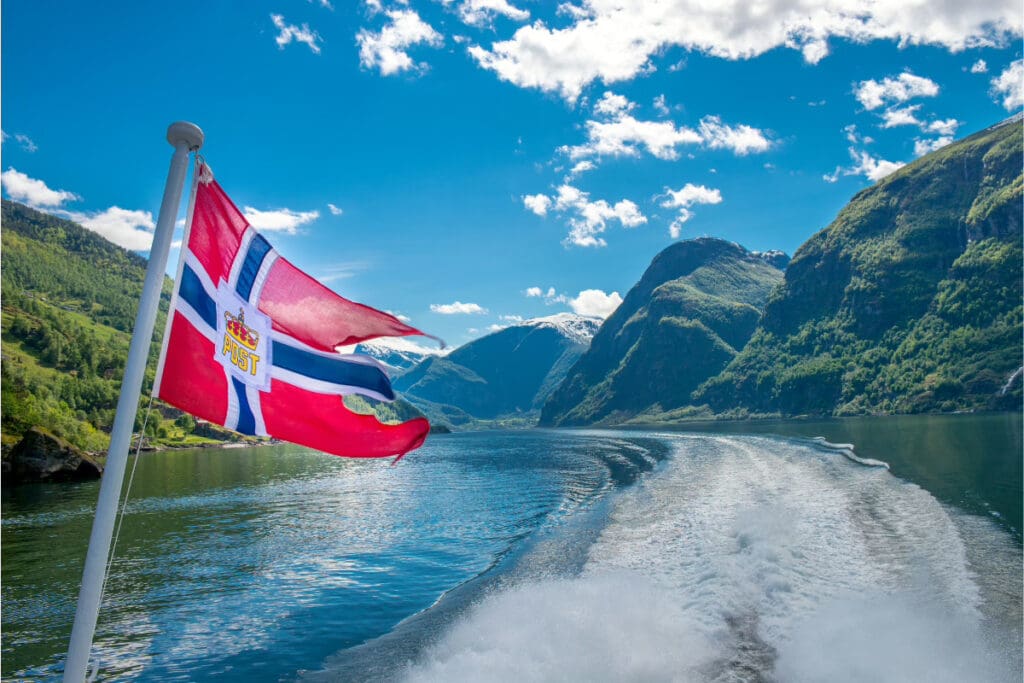 Adventure Trip Ideas, Norwegian flag waving on a boat with scenic fjord landscape in the background.