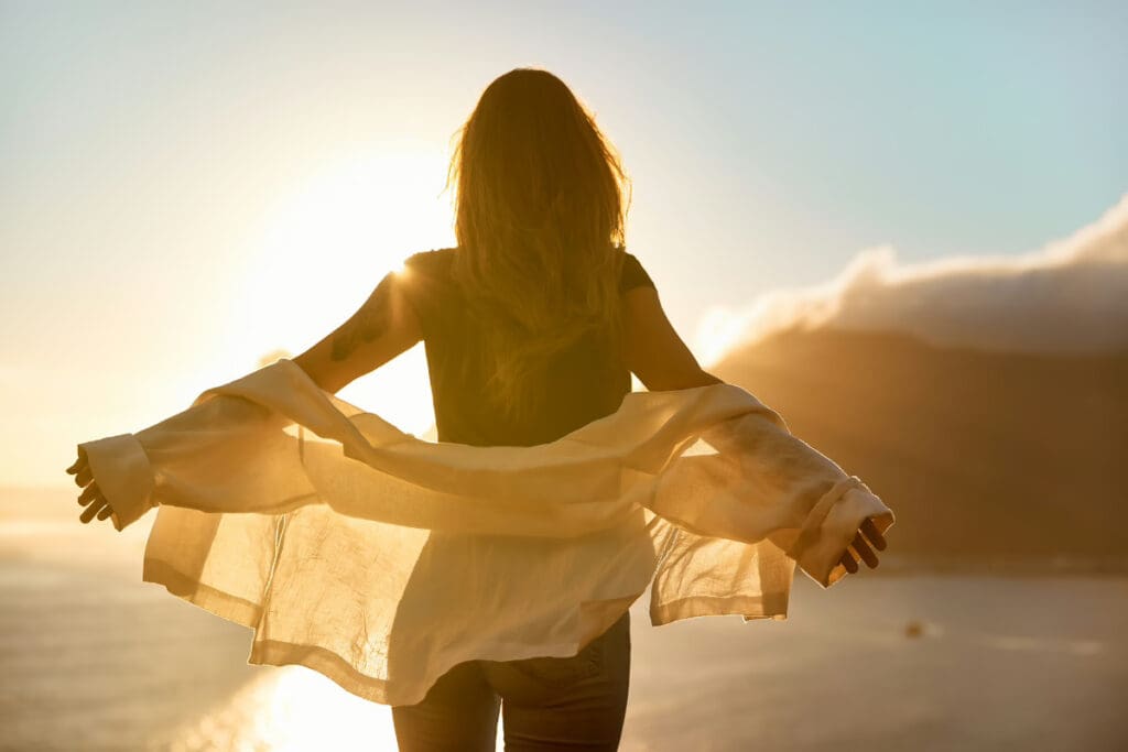 Travel on a Whim,A person with long hair standing with arms open against a sunset backdrop on a cliff.