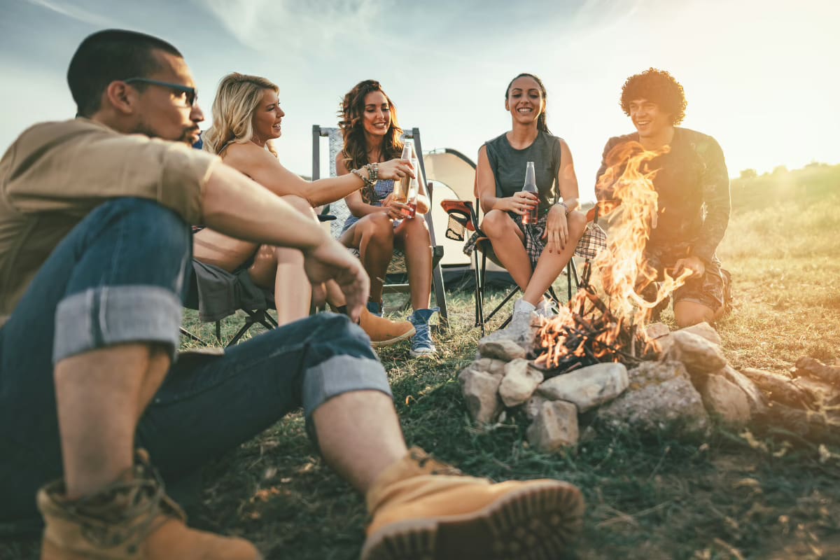 Travel on a Whim, Group of travelers celebrating around a beach bonfire during sunset with drinks in hand.