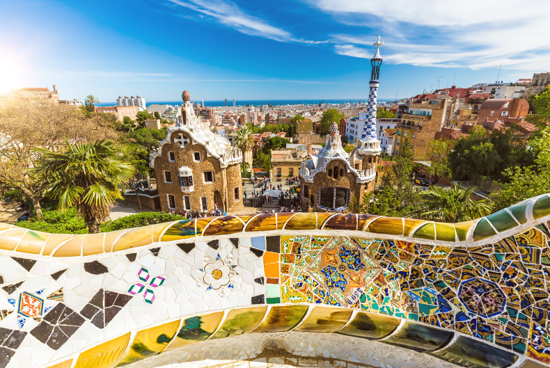 Colorful mosaic-tiled serpentine bench overlooks whimsical buildings and lush greenery in Park Güell, one of the UNESCO World Heritage Sites in Spain, with Barcelona’s cityscape and a vibrant blue sky in the background.
