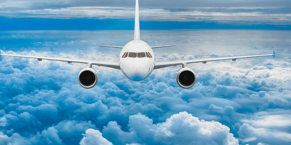 A commercial airplane flying above the clouds, facing directly toward the viewer with a blue sky and scattered cloud cover in the background.