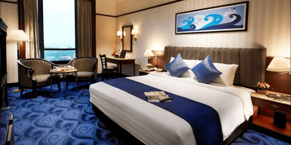 A hotel room with a large bed, blue and white bedding, two blue pillows, a table with two chairs by the window, a desk with a chair, lamps, and a framed wave artwork above the bed.