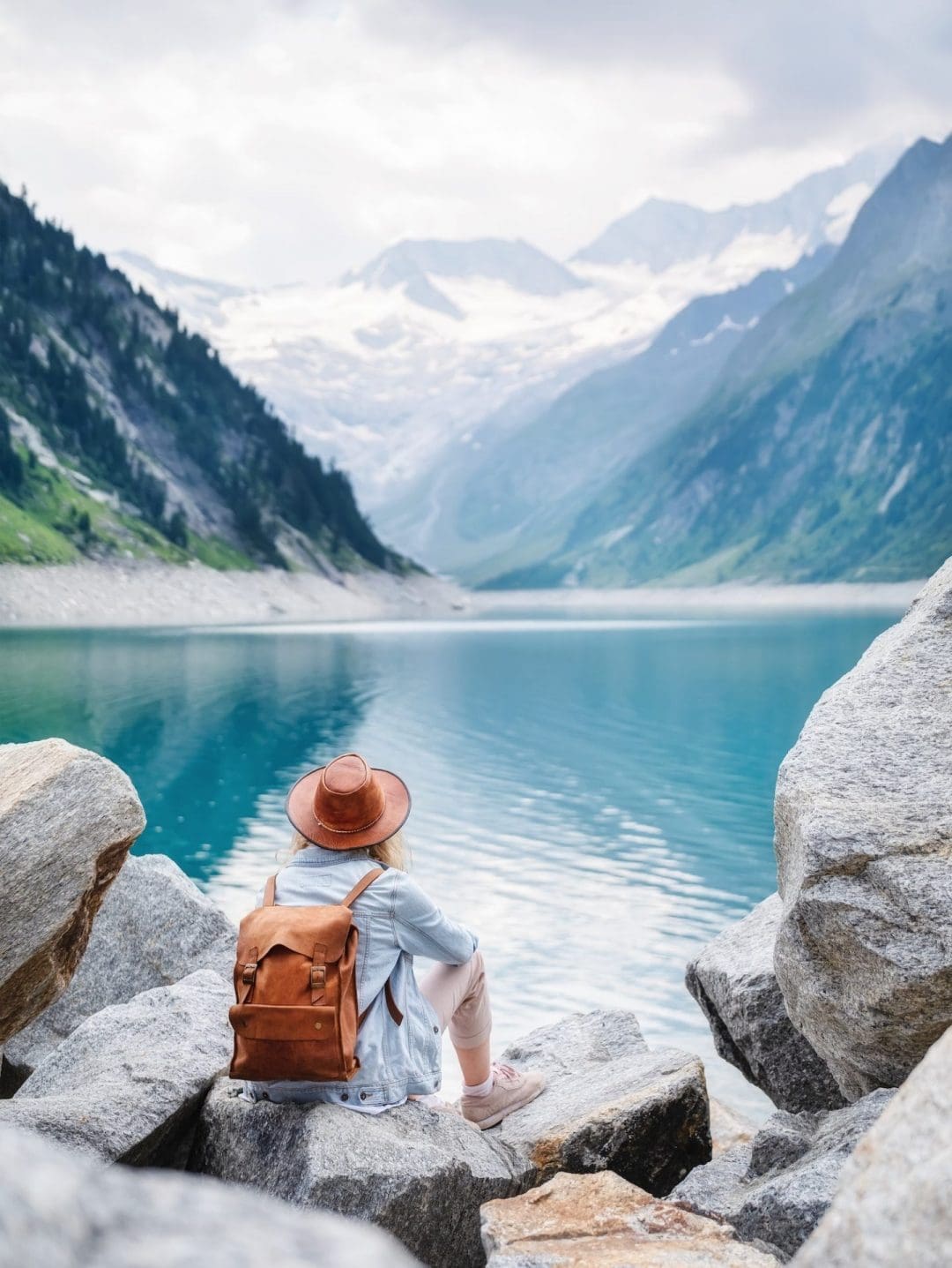 A person wearing a hat and backpack sits on rocks by a turquoise lake, surrounded by mountains with snowy peaks under a cloudy sky.