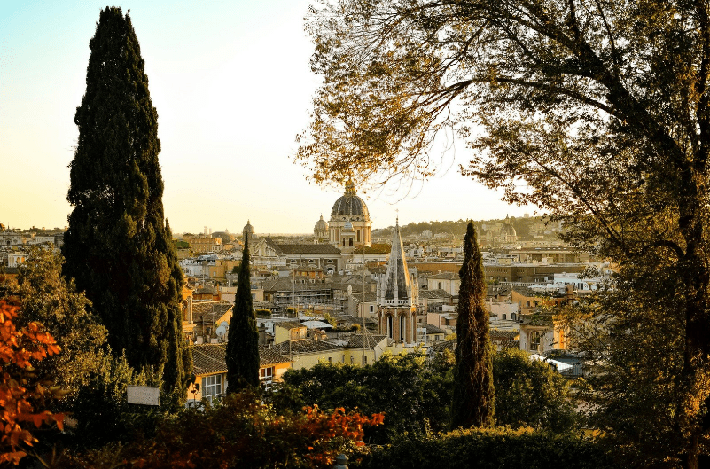 View of a city skyline with prominent domes and spires, framed by trees during sunset. HIdden gems in Rome